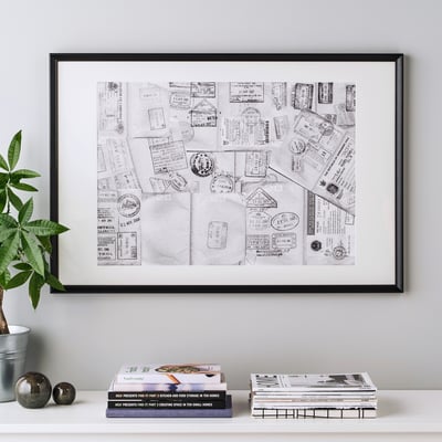 Framed travel-themed black and white art with stamps, mounted on a wall. Surrounded by books and a plant on a shelf.