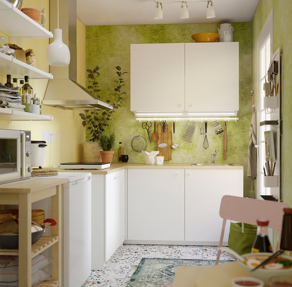 Bright kitchen with white KNOXHULT cabinets. Small, green-walled corner setup with portable hob, open shelves, and hanging utensils. 5-year guarantee.