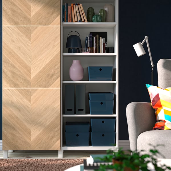 Beige cabinet with blue KUGGIS boxes and pink vase against blue wall, grey chair with colourful pillow.