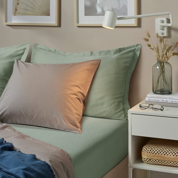 Bed with green sheet, orange pillow, grey blanket, bedside table with books, glasses, and dried flowers.