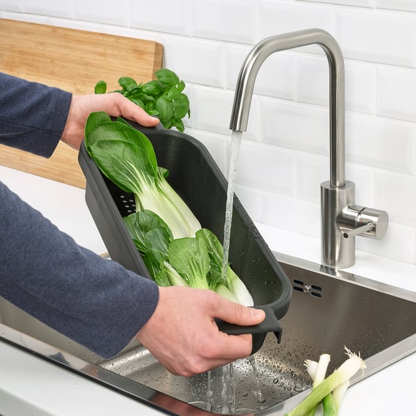 Person washes green vegetables in black LILLHAVET colander under tap over sink.