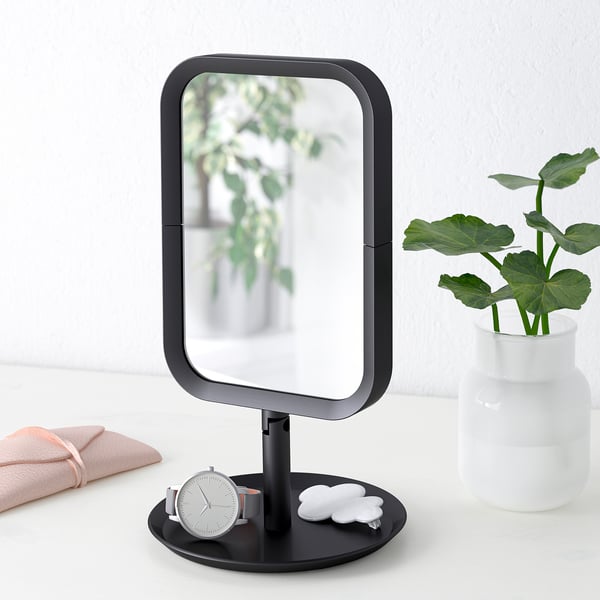 A black vanity mirror with magnified side, watch, towel, and plant on its base.