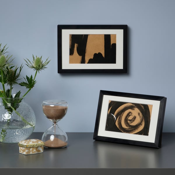Two framed artworks, one on wall, one on stand. Hourglass and vase with flowers on table. Grey wall and surface.