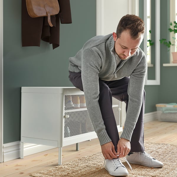 Person tying white trainers while bent over, wearing a grey sweater. Beside the person, a white storage bench with sliding doors and mesh shelves.