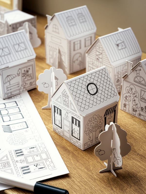 Image displays MÅLA paper craft houses, unpainted, black outlines, various shapes, on wooden table, with instructions.