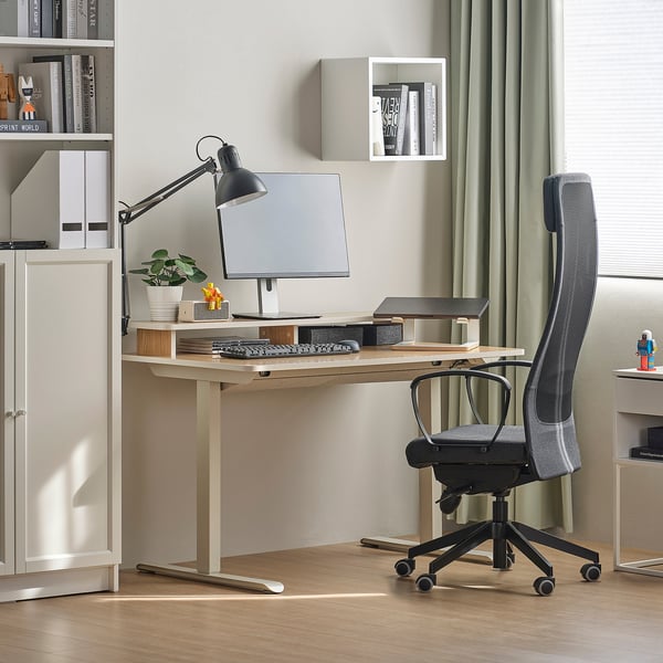 Ergonomic black office chair at wooden desk with bookshelf, monitor, and lamp.