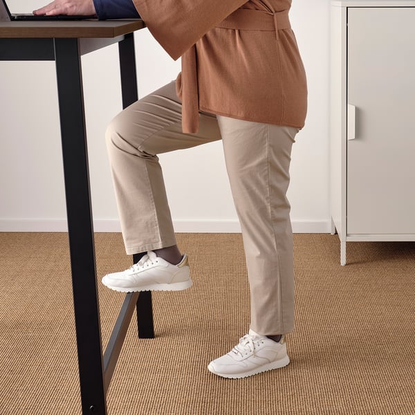 Person sitting at high table, feet resting on stool. Table has built-in power outlet and cable management. Footrest supports during long meetings.