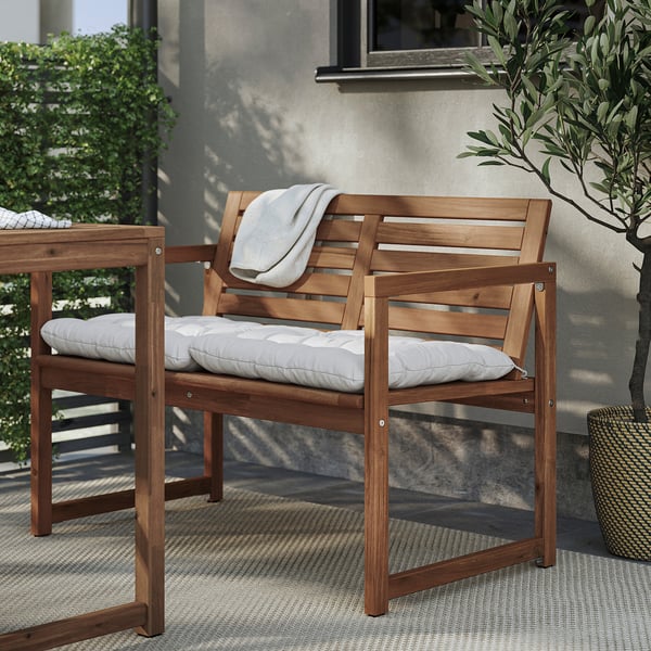 Wooden outdoor bench with light grey cushions, slatted back, placed near a house.