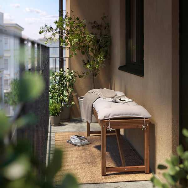 Small outdoor bench with cream cushion, wooden frame, near a building with plants.