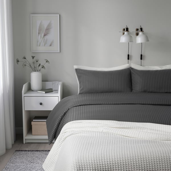 Grey bedroom with NATTJASMIN sateen duvet set and striped bedding. Neatly made bed, white accents, bedside table with dried flowers, lamp.