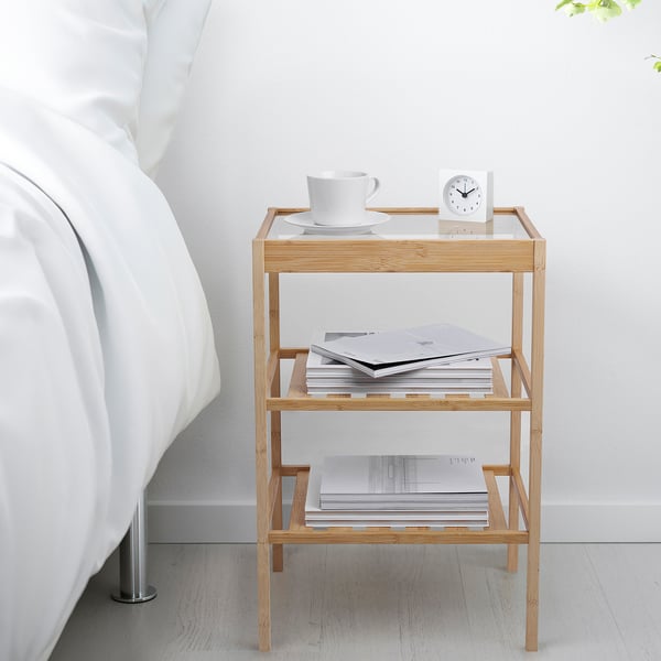 Bamboo side table with glass top & shelves beside bed, holding items.