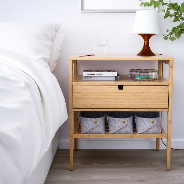 Wooden bedside table NORDKISA with drawer & shelves, holding books, lamp & bins. Light wood against white bed & grey floor.