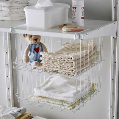 White wire basket hangs on shelf, holding folded towels. Above, filled box and small bear toy sit on shelf.