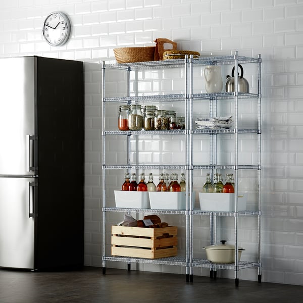 Metal shelving unit against white tiled wall, holding jars, bottles, baskets, and kitchen items, next to tall fridge.