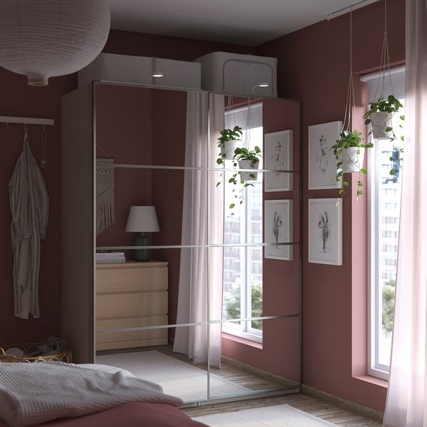 Modern bedroom with burgundy walls, featuring a reflective PAX wardrobe, hanging plants, and a window with sheer curtains.