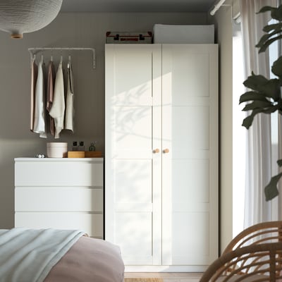 Minimalist bedroom with tall white PAX wardrobe, chest of drawers, light pink bed, and large window with sheer curtains.