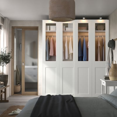 Modern bedroom with white PAX wardrobe, sliding glass doors, cosy bed, side table, warm lighting.