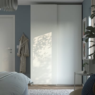 Modern bedroom with large white PAX wardrobe, hanging robe, potted plant, and bed with blue linens.