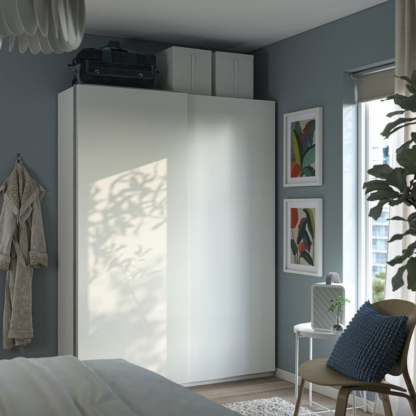 Modern blue bedroom with large white PAX wardrobe, sliding doors, and grey wall hooks. Decorated with colourful art and a cosy armchair.