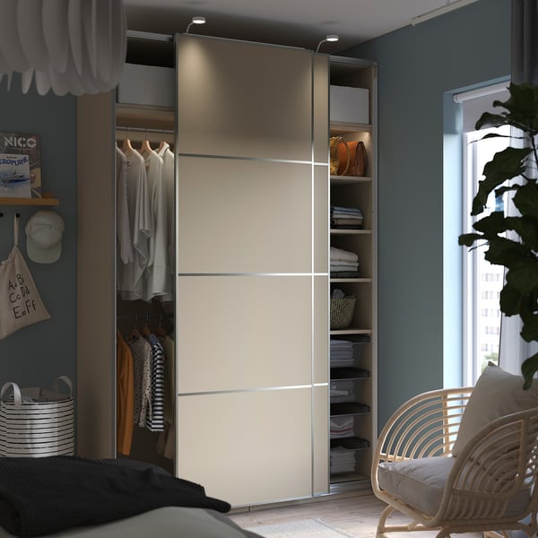 Modern bedroom with PAX wardrobe. Sliding doors reveal organised clothes and shelves. Light blue walls, wood floor, and a wicker chair.