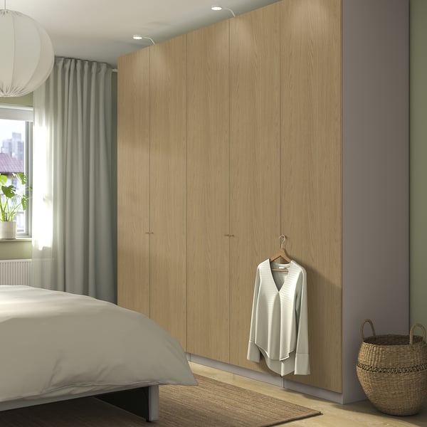 Modern minimalist bedroom with large wooden PAX wardrobe, containing light outfits. Wardrobe towers beside bed, beside wicker basket and small rug.