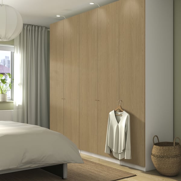 Displays a light wood-colored PAX wardrobe. Its tall and fits in a bedroom with a bed and a basket. A light sweater hangs on one door.
