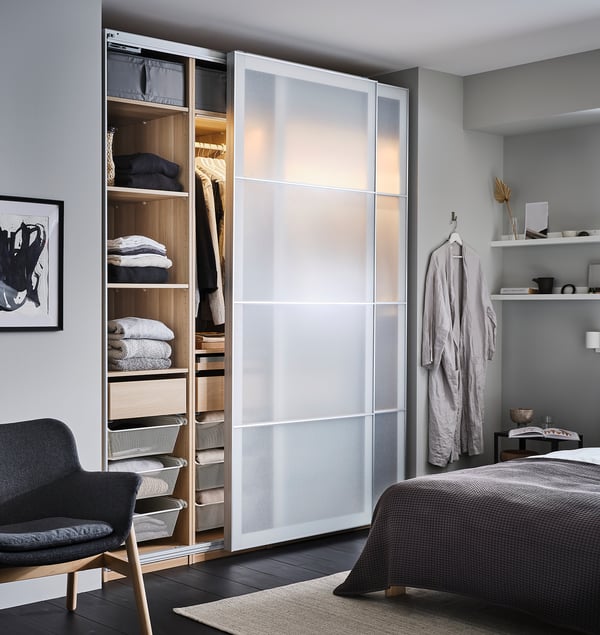 Modern bedroom with white PAX wardrobe, glass doors open to show clothes, dark grey chair, patterned bed, and decorated bedside table.