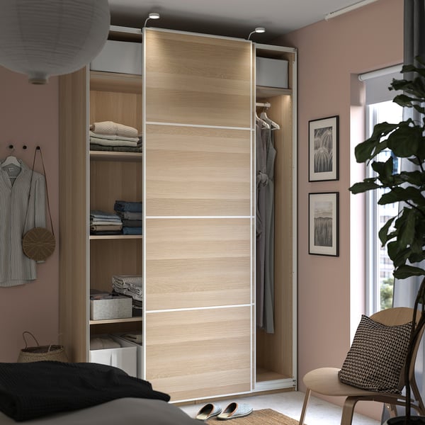Modern bedroom with PAX wardrobe in light oak. Sliding doors, shelves, and hanging racks. Decor includes striped shirts, a wicker bag, and art prints.