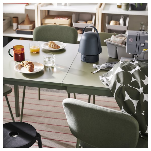 Cosy dining area with green table, chairs, and striped rug. Table has pastry, juice, mug, and lamp. Green stool and sewing machine.