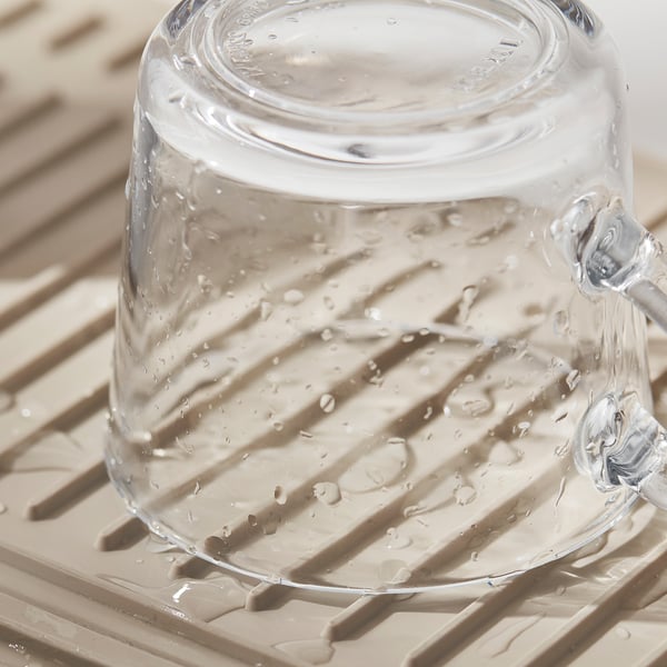 Clear glass with handles sits on a beige, ridged mat, likely a RINNIG drying mat, designed to catch water drips and keep dishes stable.