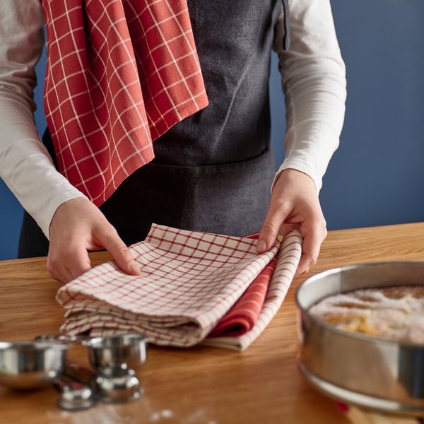 Person folding red chequered kitchen towel on wooden table with utensils and food bowl nearby.
