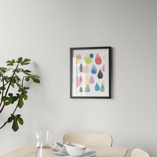 Framed colourful droplet art hangs on a wall above a wooden table with a chair and plant.