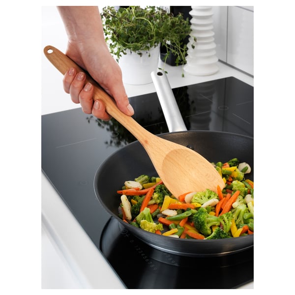 A hand holds a wooden RÖRT spoon stirring colourful vegetables in a black frying pan on a modern stovetop.