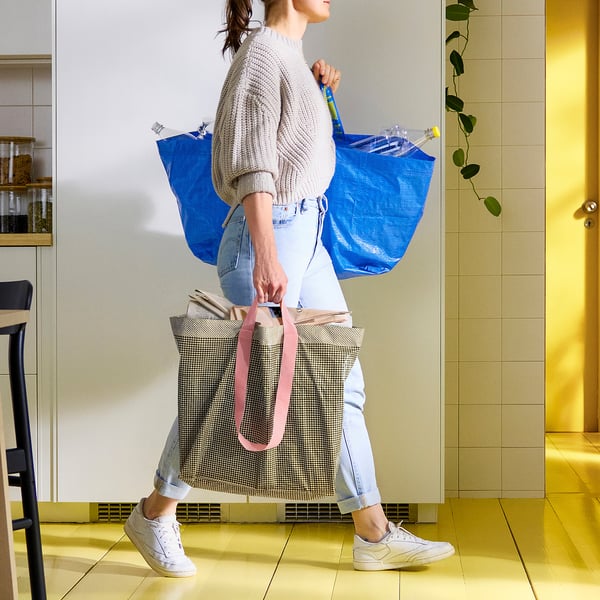 Person carrying 2 large bags: blue recycled plastic & patterned cotton tote. Standing in sunny entryway.