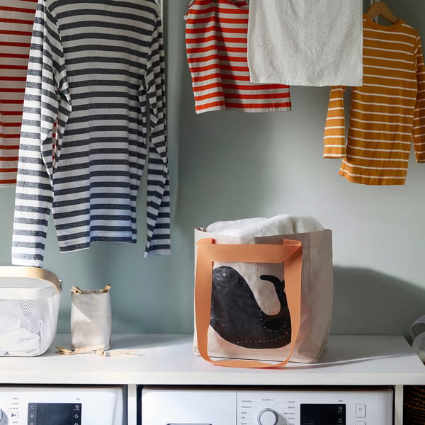Striped clothes hanging; ROMELSÖN bag on washer. Bag is peach, reusable, holds laundry.
