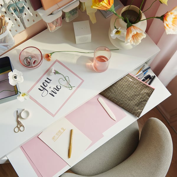 A stylish workspace with a white desk featuring a you + me card, pink accessories, flowers, and a small beige ROMELSÖN bag near notepads and pens.