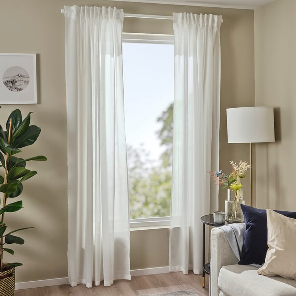 Bright living room with white curtains, chair, and lamp. Tall plant and artwork beside window.