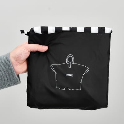Hand holding black poncho with white stripes at top, featuring a white outline of a shirt with a pocket.