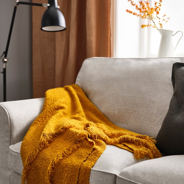 Mustard throw blanket on grey couch, soft fringed texture, with lamp and flowers nearby.