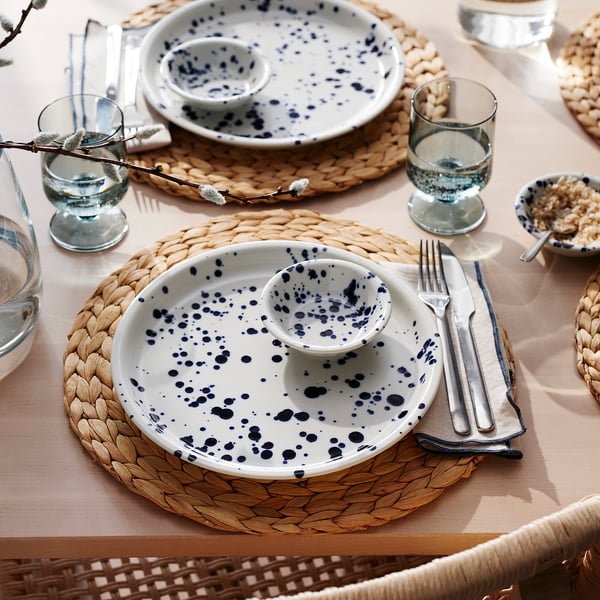 Table set with blue-speckled white plates, cutlery, glasses, and woven mats, ready for dining.