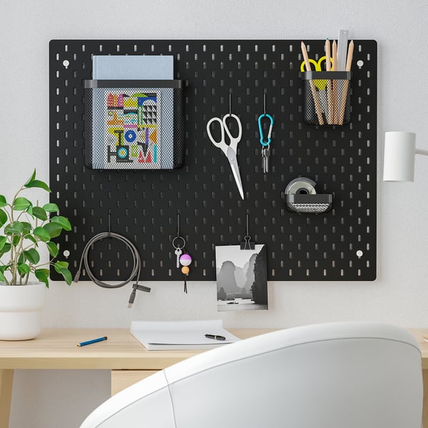 Black SKÅDIS pegboard with various accessories including a plant, pictures, and stationery hangs above a wooden desk with a white chair.