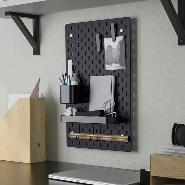 Black SKÅDIS pegboard organiser holds office supplies, notes, and accessories. Mounted on wall with shelf and hooks for storage.