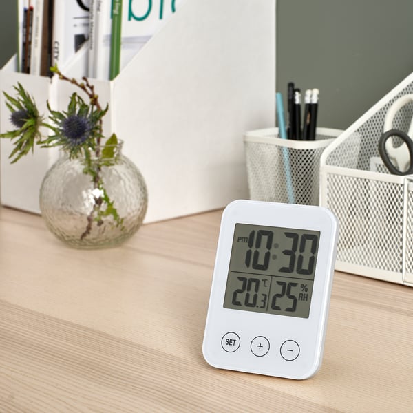 Desk scene with white SLÅTTIS multi-function clock showing time, temperature, humidity. Includes small decorative plant & pens. Clock is compact.