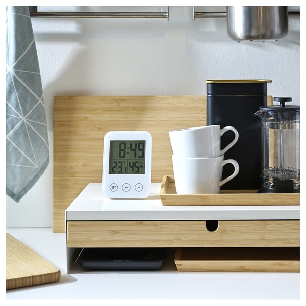 Modern kitchen scene with a white digital clock, stacked mugs, and a coffee press on a counter.