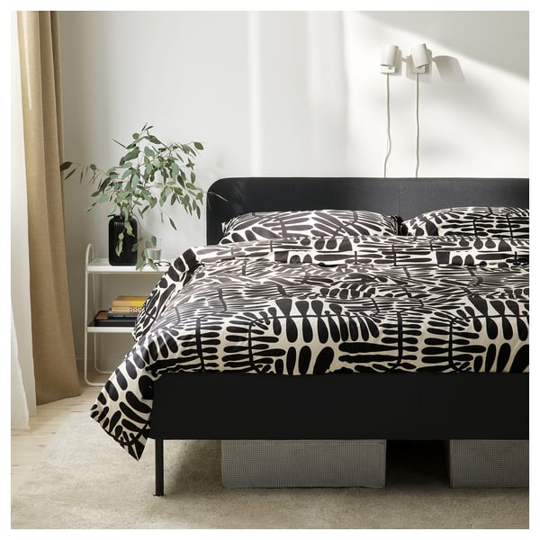 Modern black upholstered bed with padded headboard, abstract print duvet cover, stylish bedside table with plant, beige curtains.