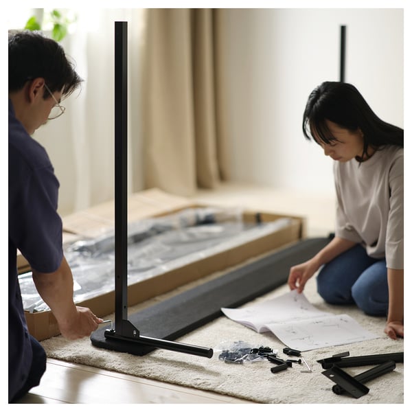Two people assemble a flat-packed SLATTUM bed frame; dark grey, modern design with upholstered headboard and steel slats.