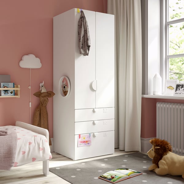 Small, white kids wardrobe with drawers, mirror, and hooks stands beside a bed, near a window. A rug and stuffed animals are on the floor.