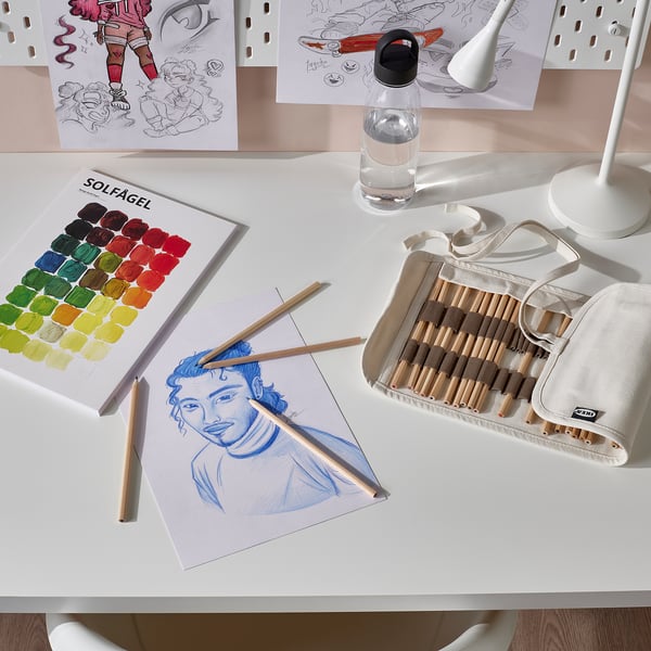 Art station setup with SOLFÅGEL coloured pencils, sketchbook, pencil case, water bottle, and drawings on white desk.