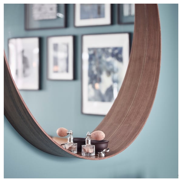 A walnut-framed round mirror with a shelf at the bottom for small items, leaning against a blue wall with framed pictures.