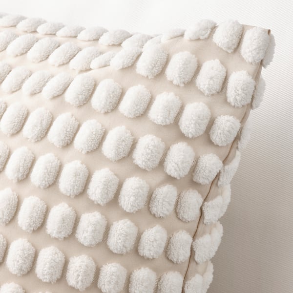 A close-up of a soft, textured cushion with white pom-poms on an off-white base. Details include a zip for easy removal.